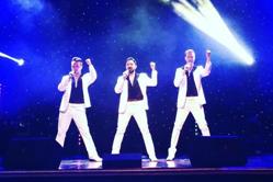 Three male vocalists performing on stage in coordinated white outfits under dramatic concert lighting.