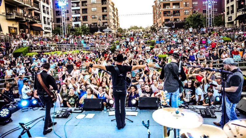 Large outdoor concert with live band performing on stage before a packed crowd.
