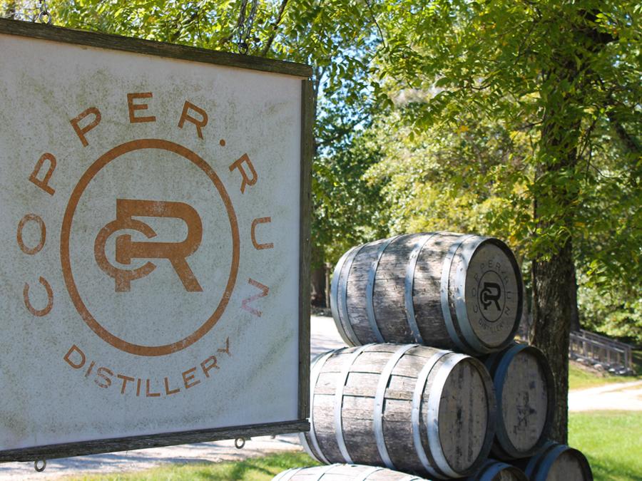 Copper Run Distillery sign beside stacked oak barrels surrounded by trees, offering visitors unique local flavor.