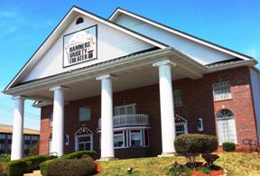 Brick building with white columns and landscaped grounds under a clear blue sky.
