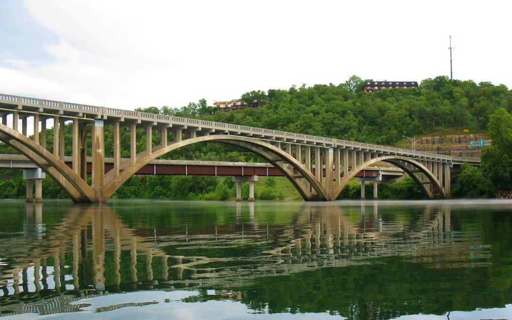 Scenic arched bridge spanning calm waters with forested hills in the background.