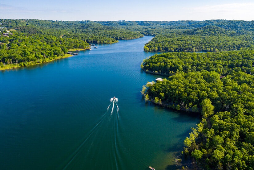 Aerial view of the winding lake surrounded by dense green forest with boats cutting through calm water.