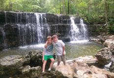 Visitors standing on rocks beside a cascading Ozark waterfall surrounded by forest.